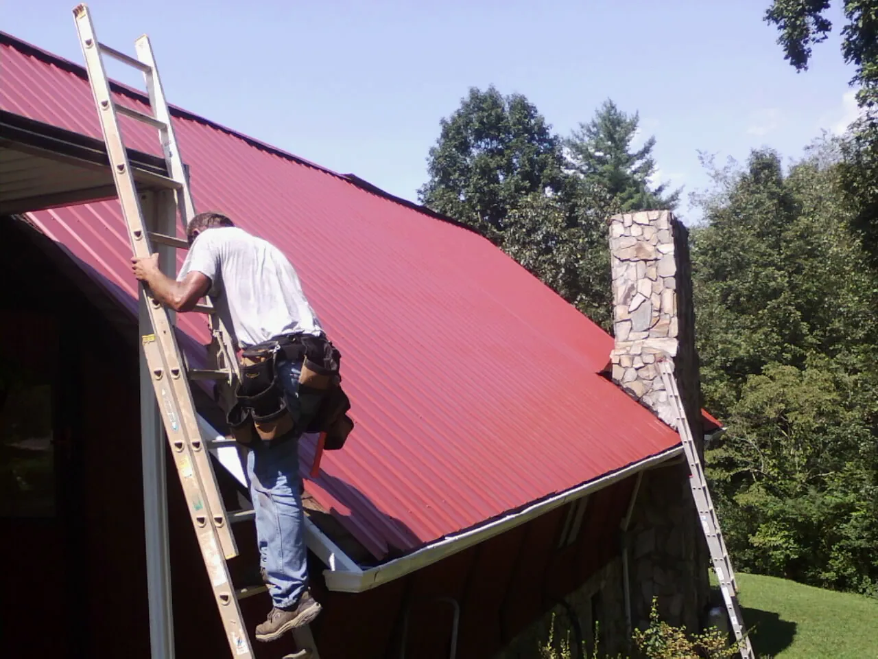 Roofer on a ladder beside a red metal roof and stone chimney — Soffit & Fascia Repair by Roofing Brothers in Valley City